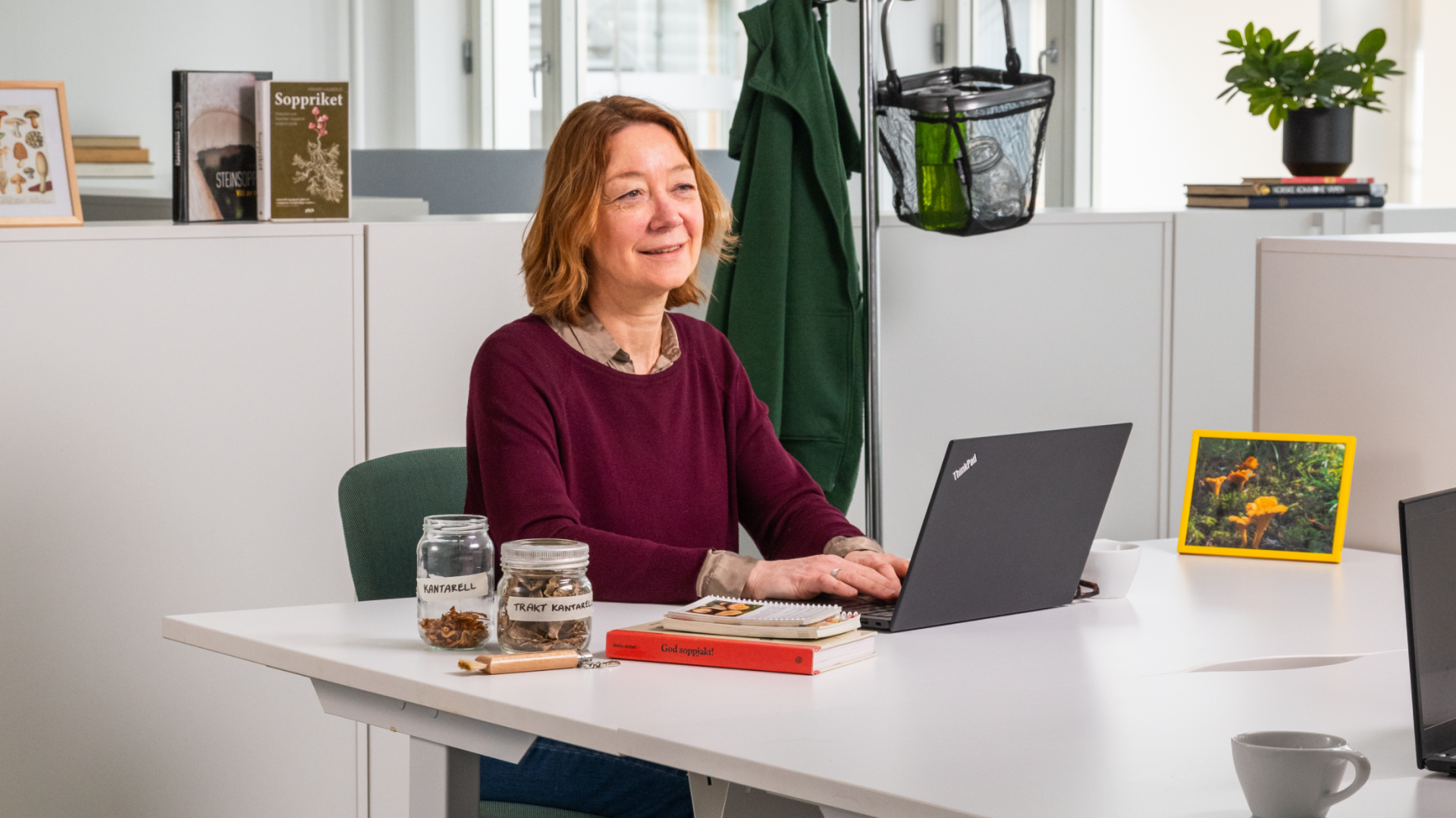 En dame sitter i et kontorlandskap foran en PC og ser tankefullt ut i luften. Rundt henne ligger soppbøker og tørket sopp